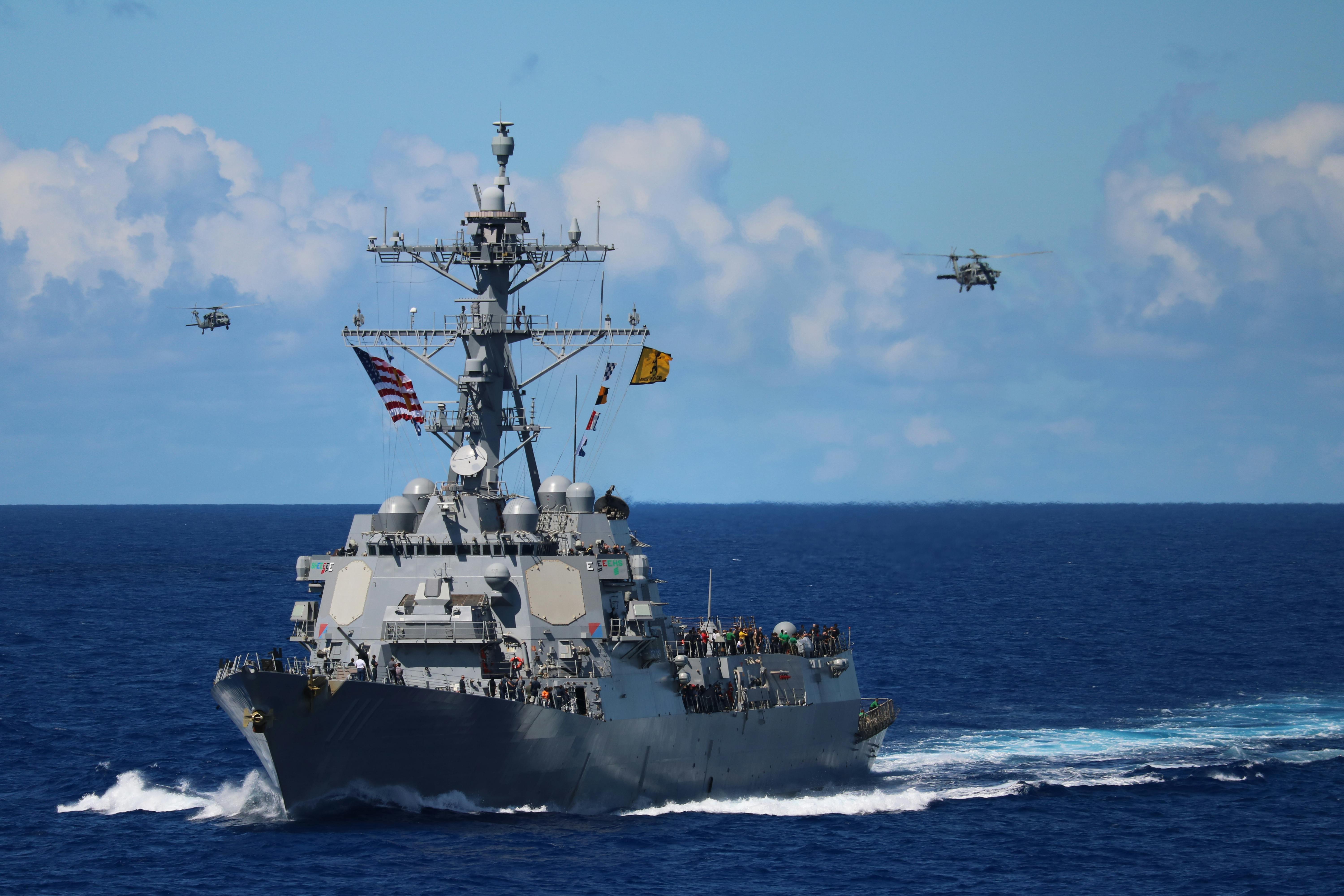 A US Navy destroyer navigates the Strait of Hormuz with helicopters in formation above.