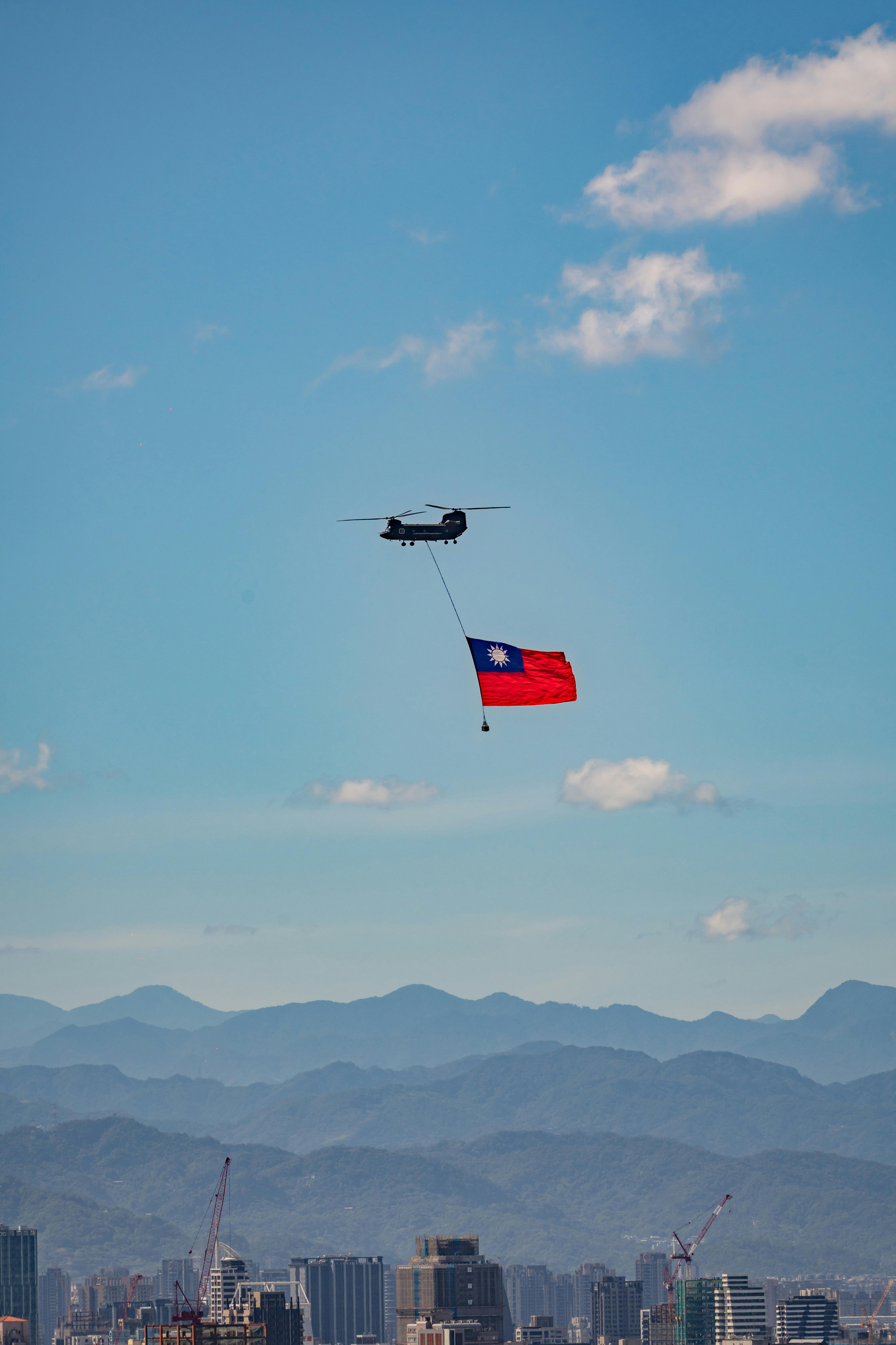Helicopter flying over Taipei skyline carrying Taiwanese flag against mountainous backdrop