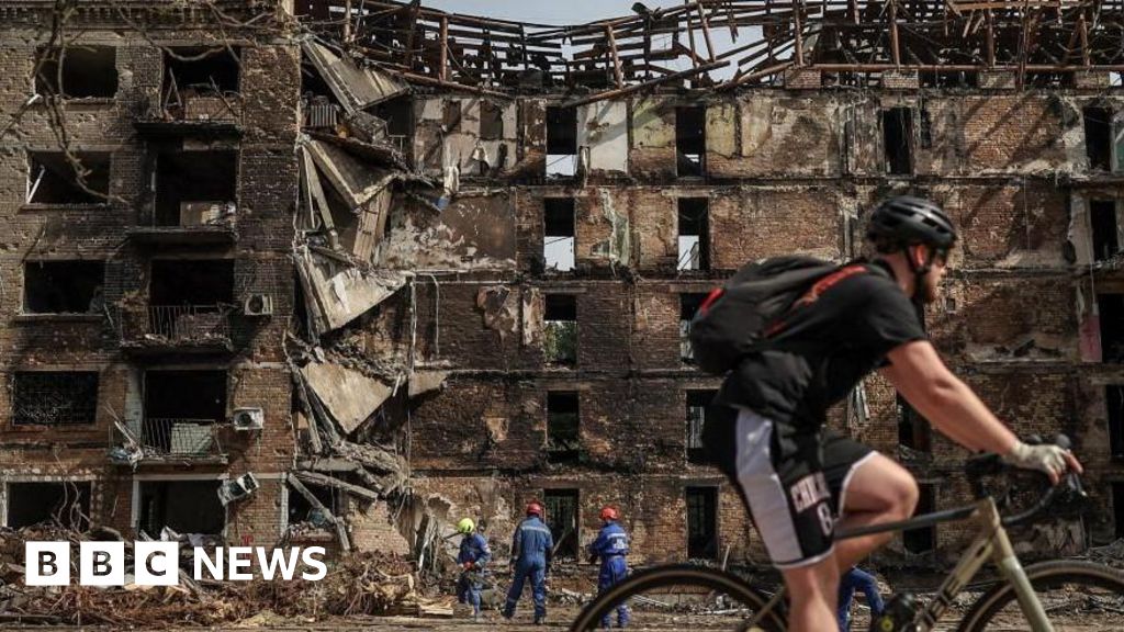 A cyclist rides past a damaged building, with a crew cleaning up in front of it